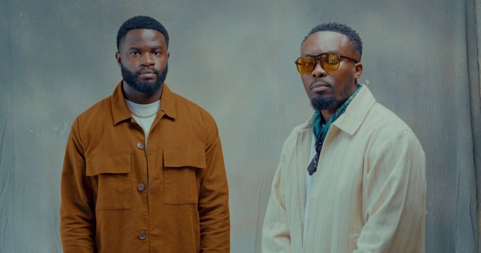 Faith Child (right) and Marizu Ikechi (left), standing calmly side by side for the single, "Oluwa is Involved." Faith Child is wearing a cream shirt with black stripes and a different, brown tinted glasses, while Marizu is wearing a burnt orange shirt with a white t-shirt underneath it.