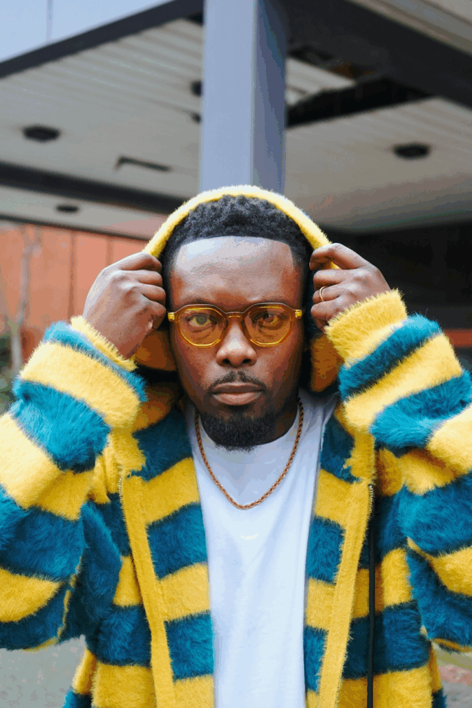 One of U.K.'s best contemporary artists, Faith Child, is wearing a fuzzy yellow and blue big-striped hoodie with brown tinted glasses and a white t-shirt underneath the hoodie. Faith Child is holding the top of the hoodie over his head as if he were just to be wearing it.